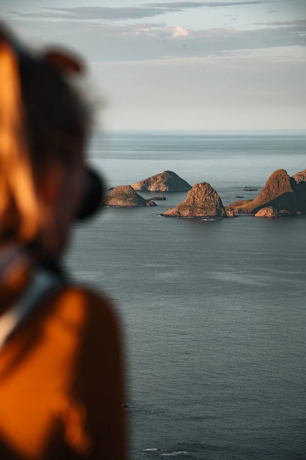 Annina unscharf mit der Kamera im Vordergrund, dahinter eine Gruppe Felsinseln im Meer, vom Skårvågsfjellet aus