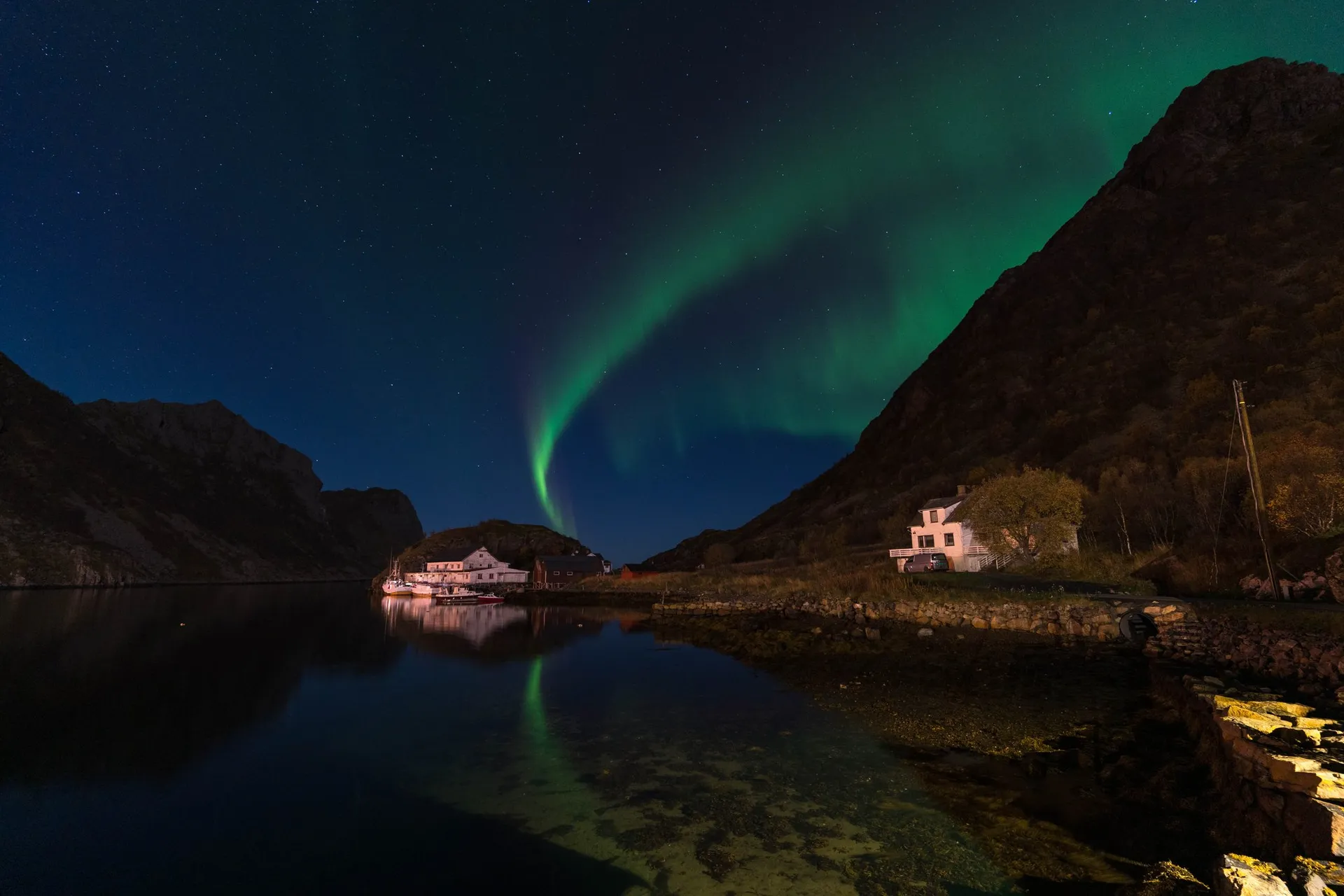 Ein grüner Nordlichtbogen am Sternenhimmel über einer Bucht mit Häusern und Booten, das Licht spiegelt sich im stillen Wasser