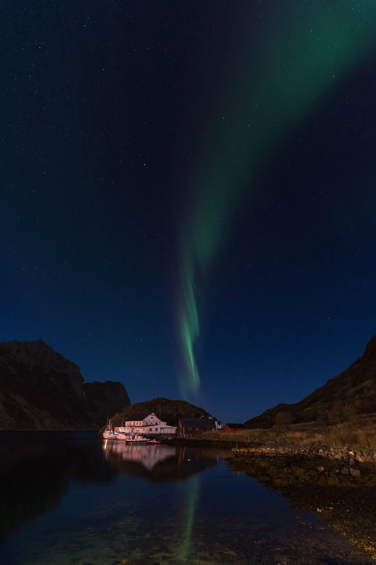Ein grünes Nordlichtband zieht senkrecht über den Sternenhimmel, darunter Häuser am Ufer einer stillen Bucht
