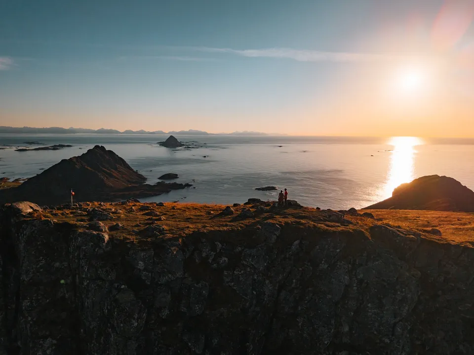 Zwei kleine Figuren am Rand des Skårvågsfjellet im Abendlicht, links der spitze Gullstabben, in der Mitte die Insel Gaukværøya, am Horizont die Lofoten