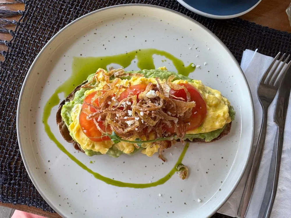 Toast with avocado, scrambled egg, tomato slices and crispy strips of onion on a light plate with dark speckles