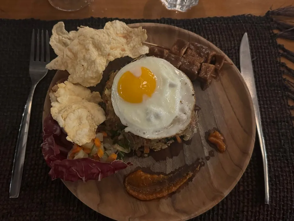 Nasi Goreng mit Spiegelei auf einem Holzteller, daneben zwei helle Cracker, Gemüsewürfel und ein Spiesschen
