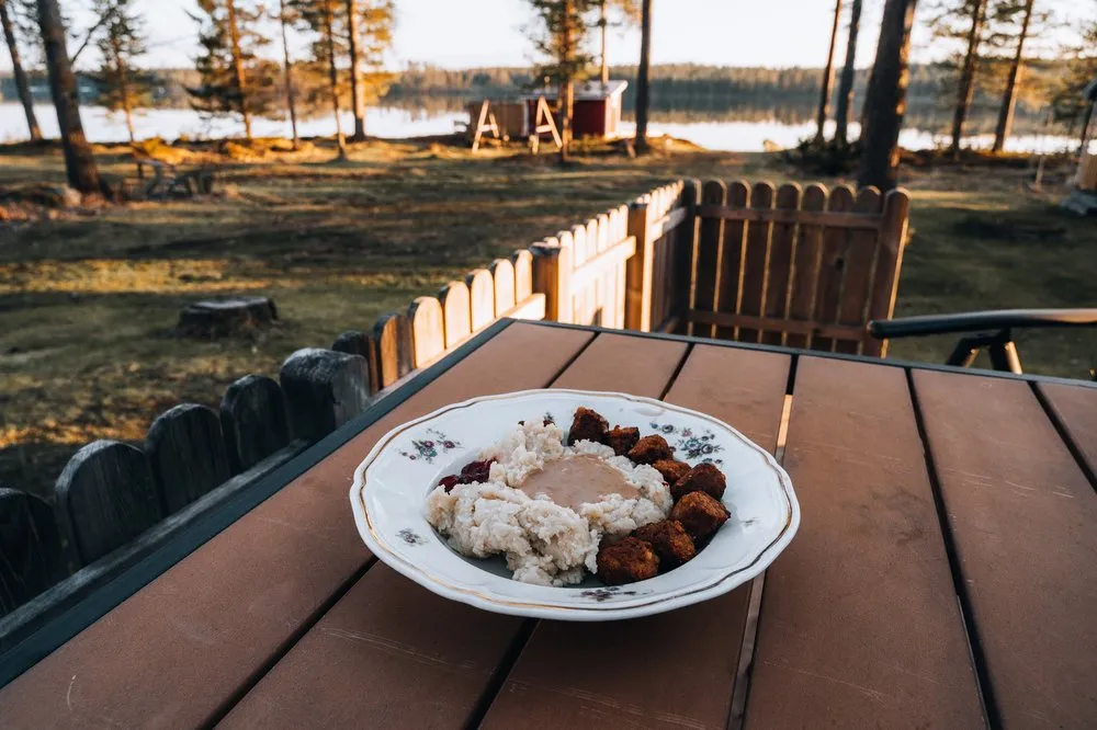Teller mit Kartoffelstock, Sosse und Köttbullar auf dem Terrassentisch, dahinter Wiese und der See im Gegenlicht