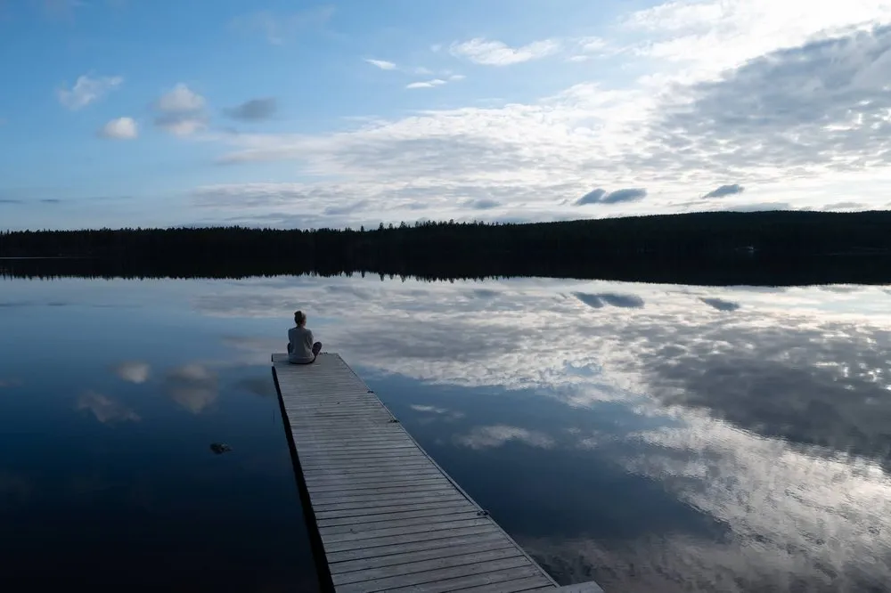 Eine Person sitzt am Ende des Stegs, Himmel und Wolken spiegeln sich im dunklen See