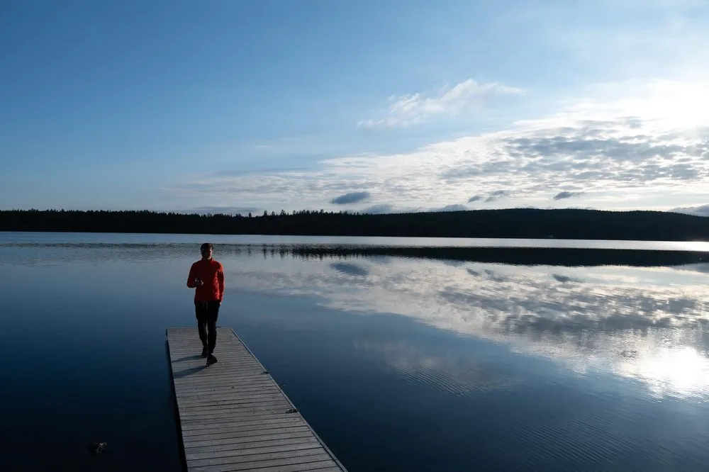 Ein Mann in roter Jacke steht auf dem Steg, dahinter blauer Himmel und der ruhige See