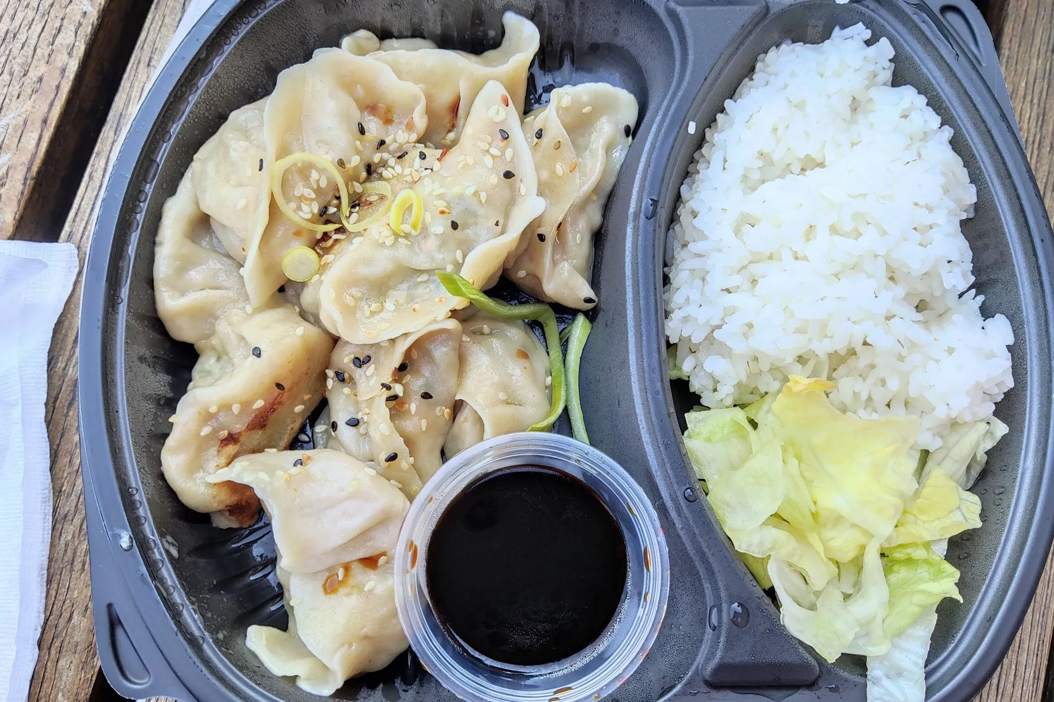 From above: Dumplings with soy sauce, rice and salad leaves in a black takeaway container on a wooden bench