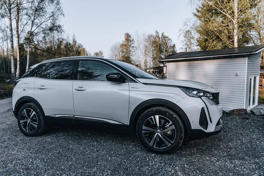 Weisser SUV auf einem Kiesplatz zwischen Birken und Nadelbäumen, rechts ein graues Holzhäuschen