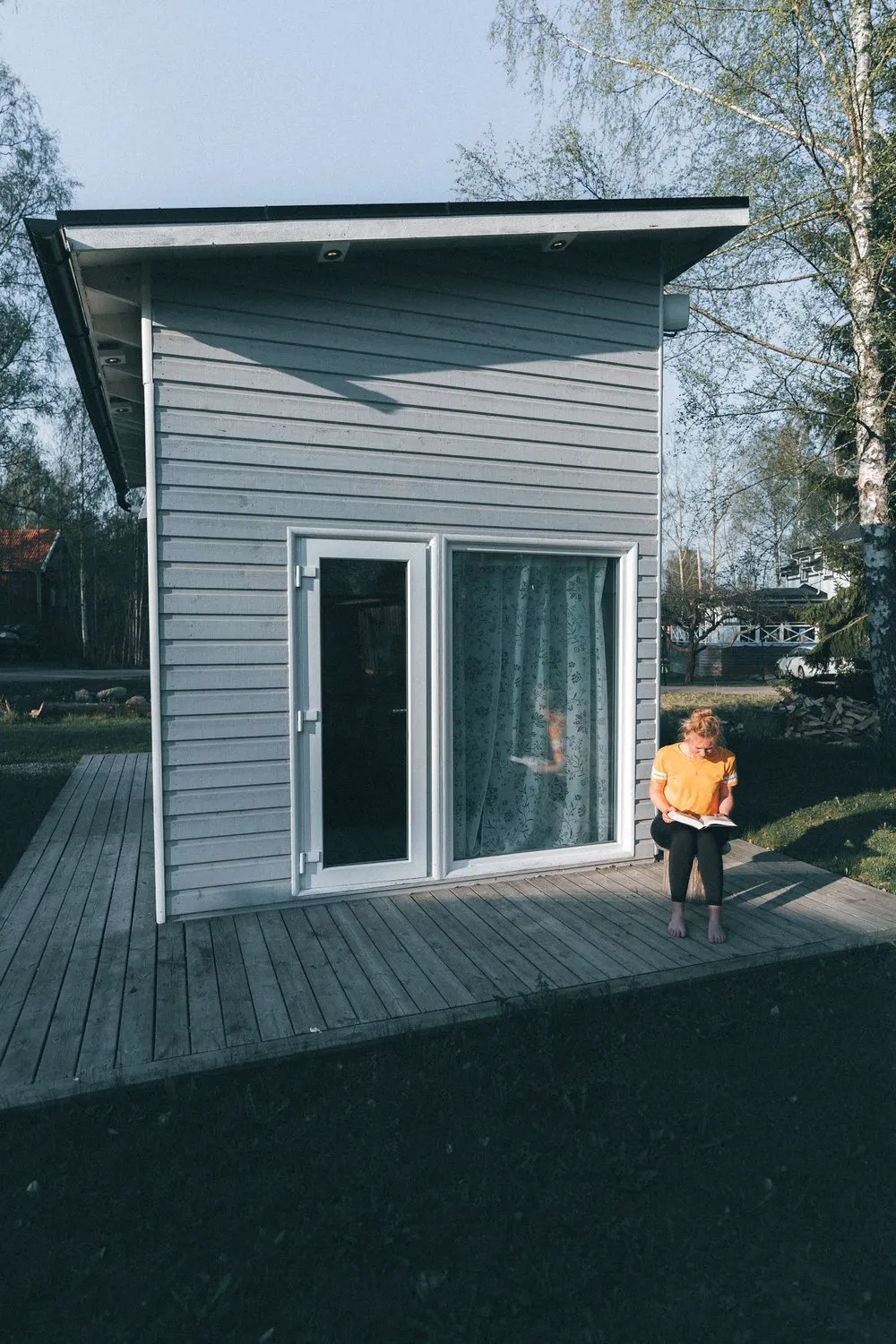 Das Tinyhouse mit grauer Holzverkleidung und Terrasse, davor sitzt eine Person mit orangem Oberteil und liest