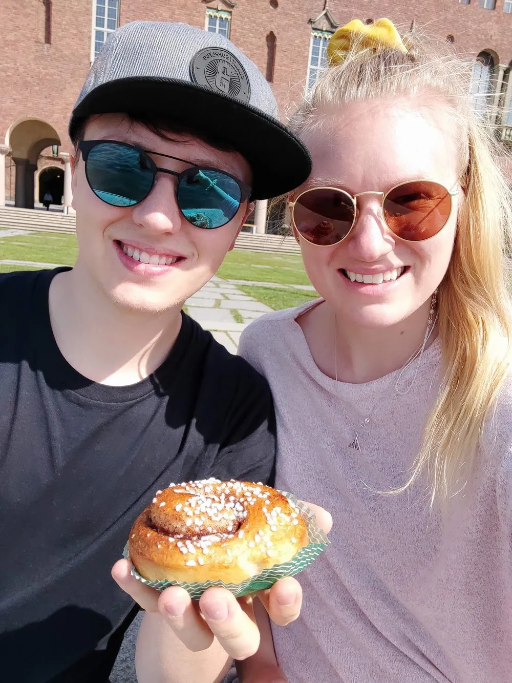 Two people in sunglasses holding a cinnamon bun up to the camera for dessert at the Stadhus, a brick facade with round arches behind them