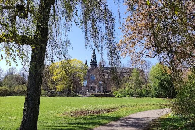 Wiese und Spazierweg im Park auf der Insel, dahinter ein Backsteingebäude mit Turm zwischen Bäumen im frischen Frühlingslaub