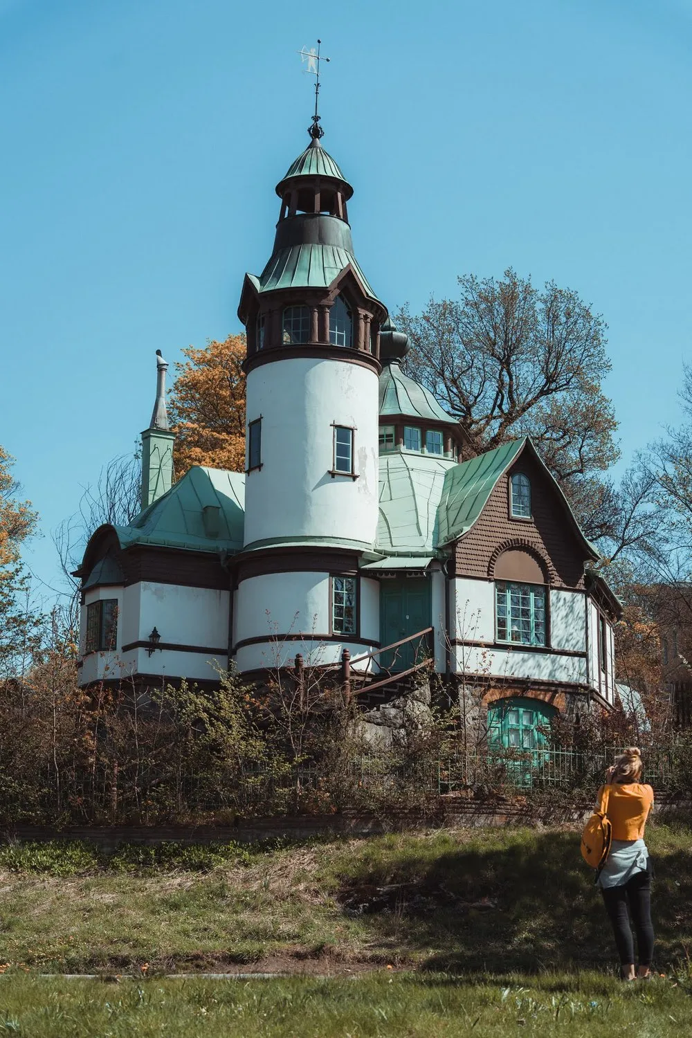 Villa mit weissem Rundturm, grünem Kupferdach und Holzverkleidung, davor im Gras eine Person mit orangem Oberteil beim Fotografieren
