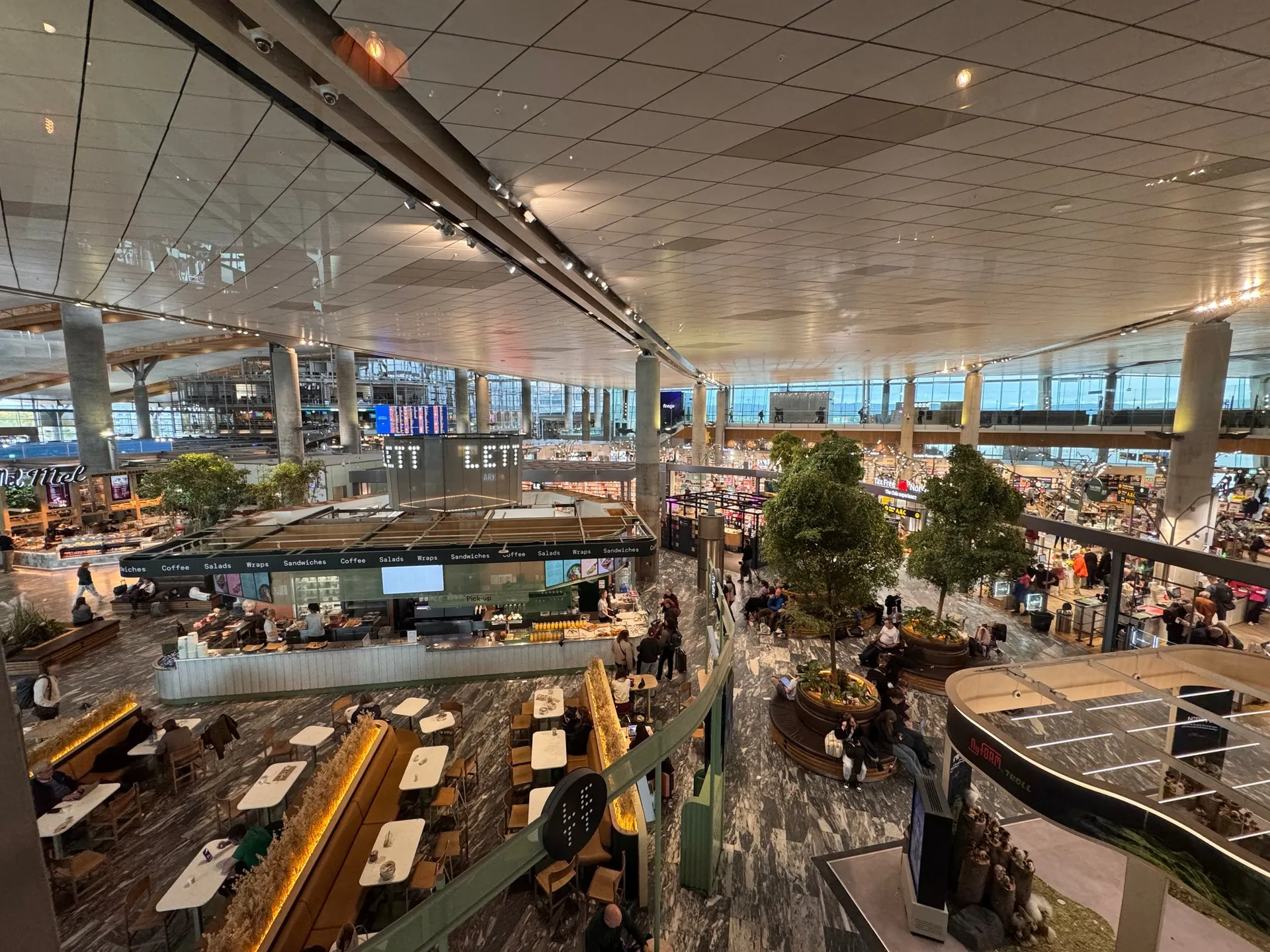 Blick von der Trattoria Tavolare hinunter in die Halle des Flughafens Oslo-Gardermoen mit Restaurants, Sitzplätzen und Bäumen
