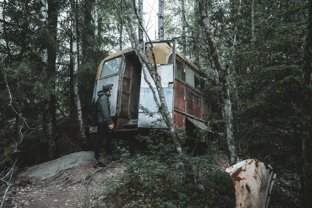 A man in a cap with a backpack stands on a rock in front of a rusty caravan in the forest