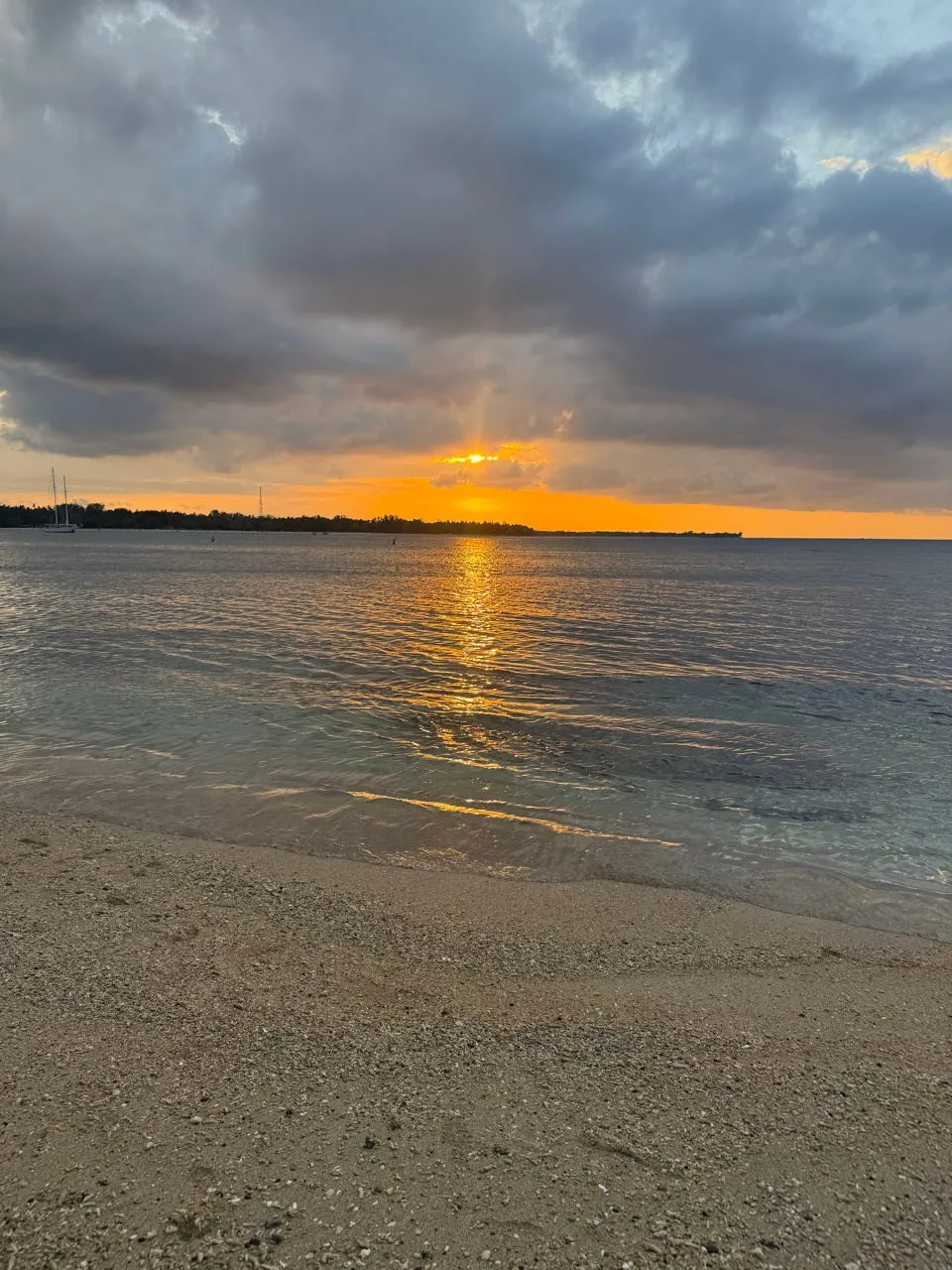 Die Sonne steht orange unter einer dunklen Wolkendecke über dem Meer, im Vordergrund nasser Sand