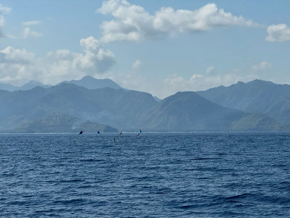 Blue mountain ranges above the sea, with a few small boats with dark sails in front