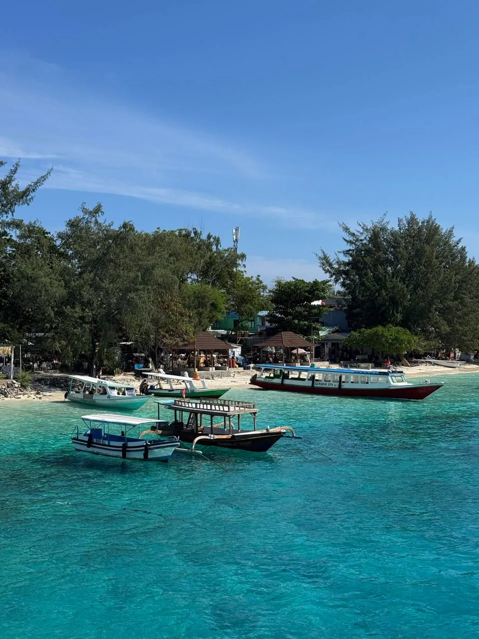 Türkisfarbenes Wasser vor einem Strand mit Booten, Bäumen und kleinen Pavillons