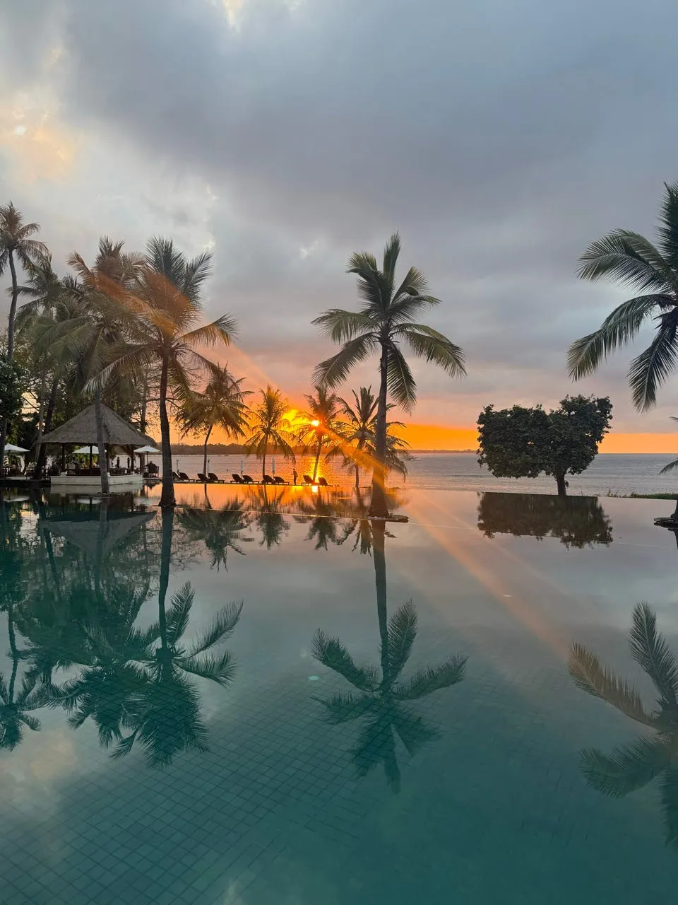 Sonnenuntergang über dem Infinity-Pool, Palmen spiegeln sich im Wasser, links ein Pavillon mit Strohdach
