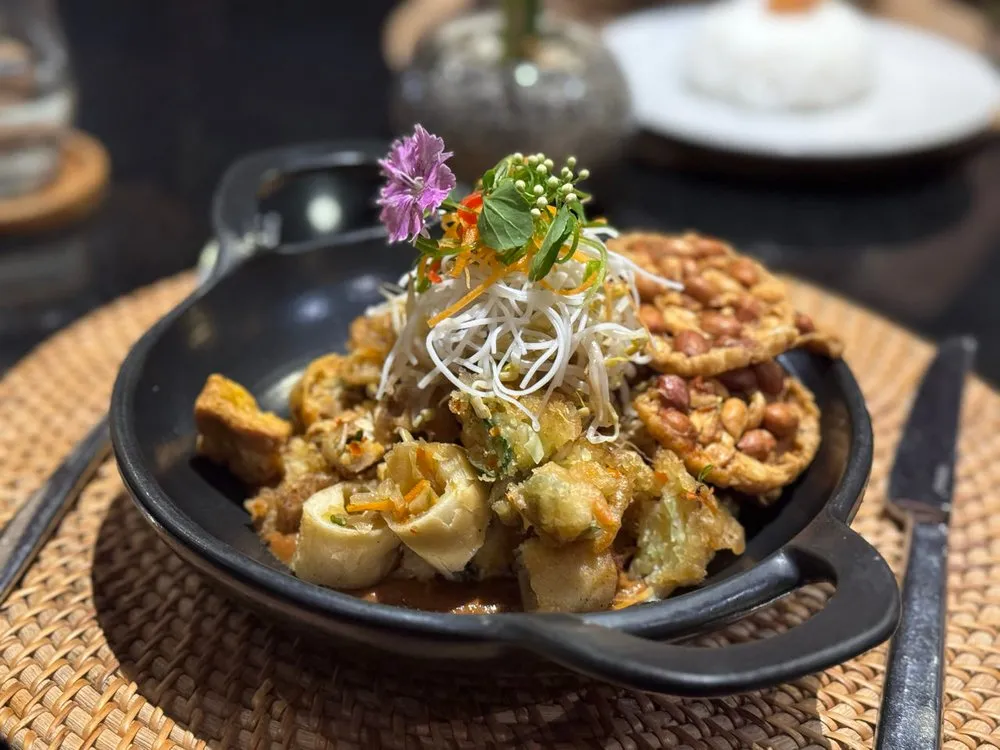 A black pan with pieces in brown sauce, topped with fine white strips and a purple flower