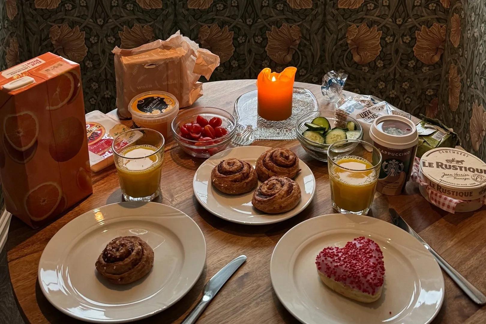 A breakfast table with a candle, cinnamon buns, cherry tomatoes and orange juice