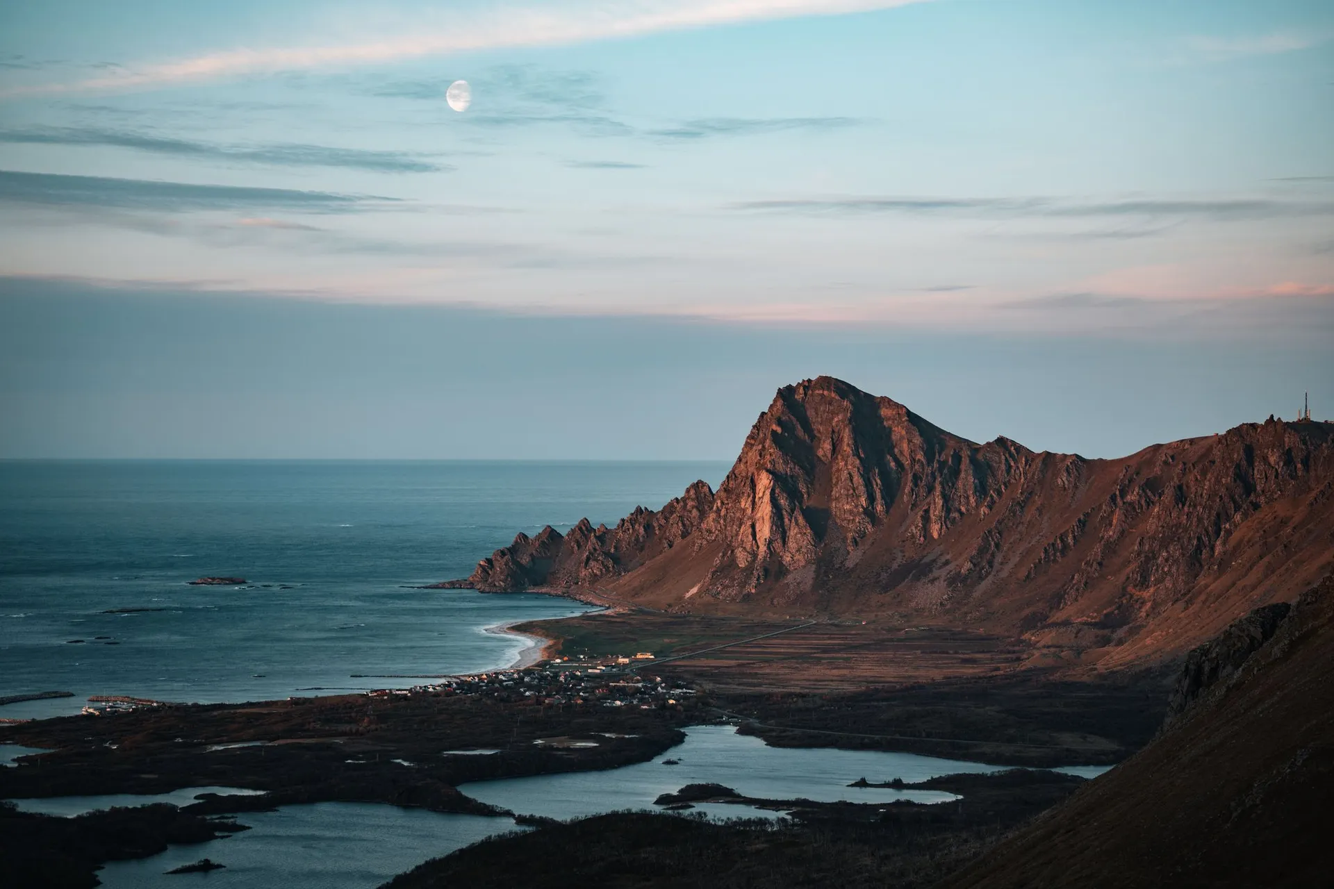 Der Mond steht über einem Berg im Abendlicht, darunter ein Dorf, Seen und das Meer, beim Abstieg vom Måtind