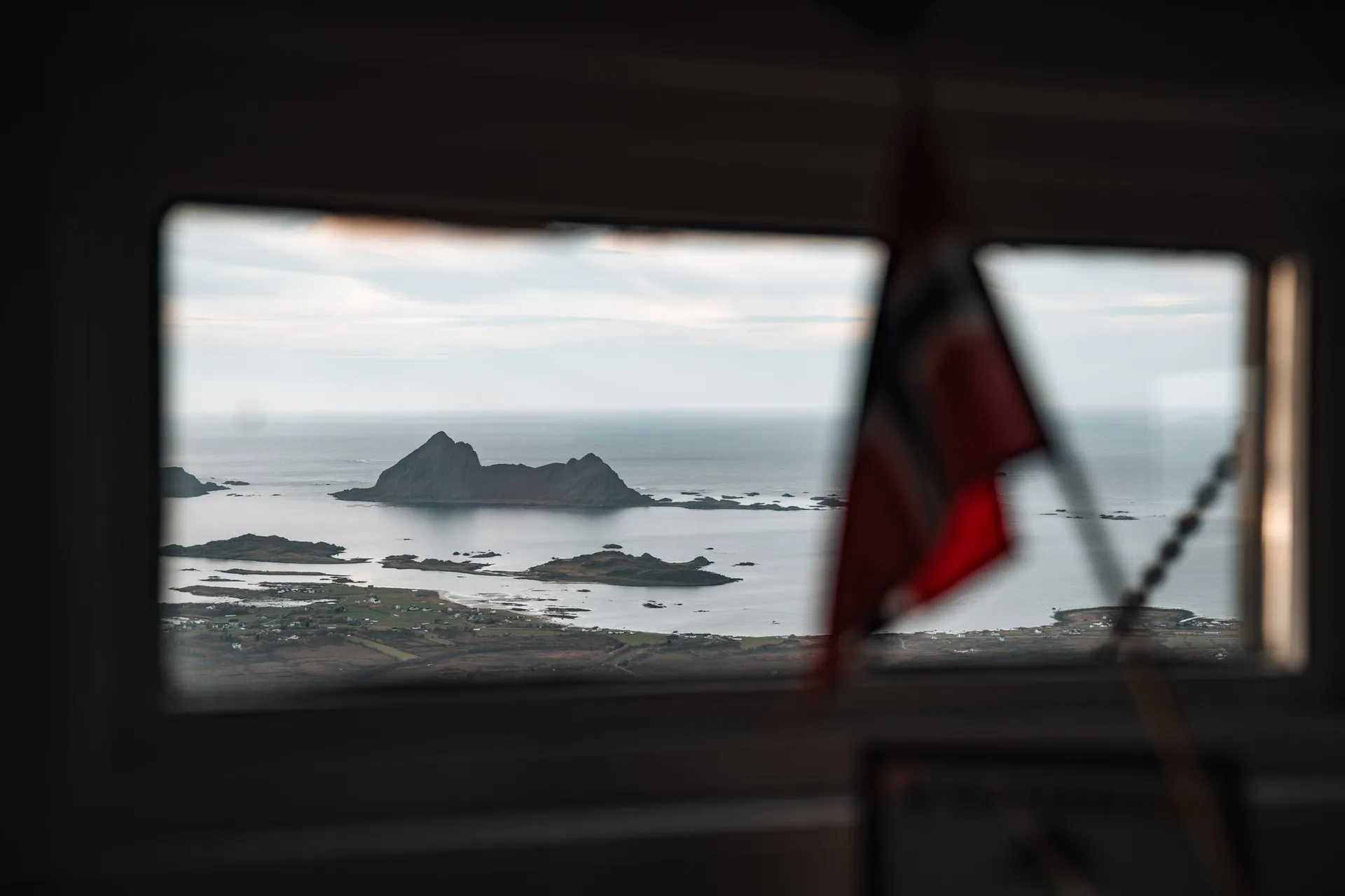 Durch das Fenster der Vettenbua auf das Meer und eine Felsinsel, vorne unscharf die norwegische Fahne