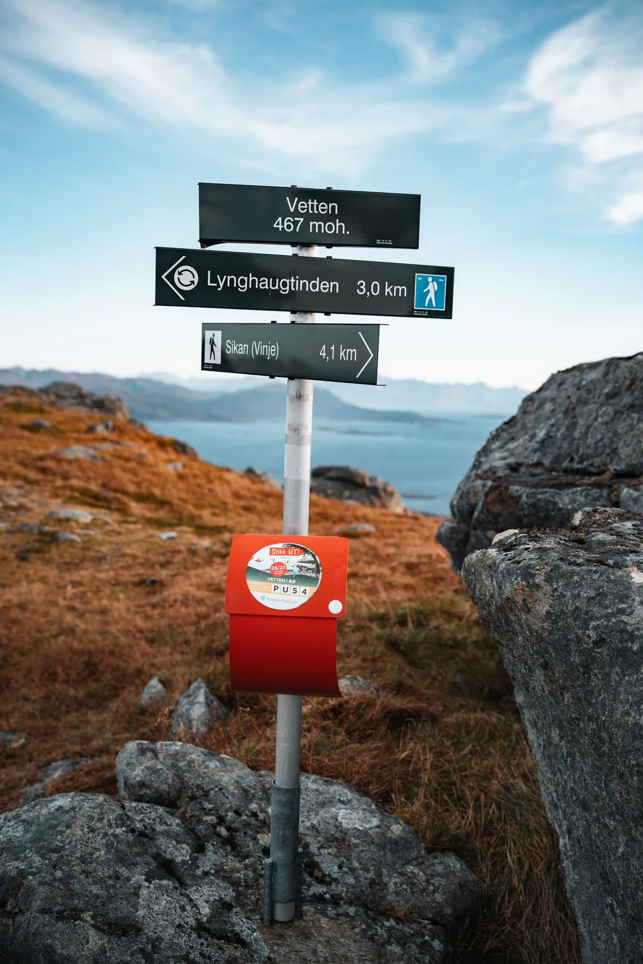 Der Wegweiser «Vetten 467 moh.» mit einem roten Kasten auf dem Gipfel, daneben Felsen und Herbstgras, dahinter das Meer