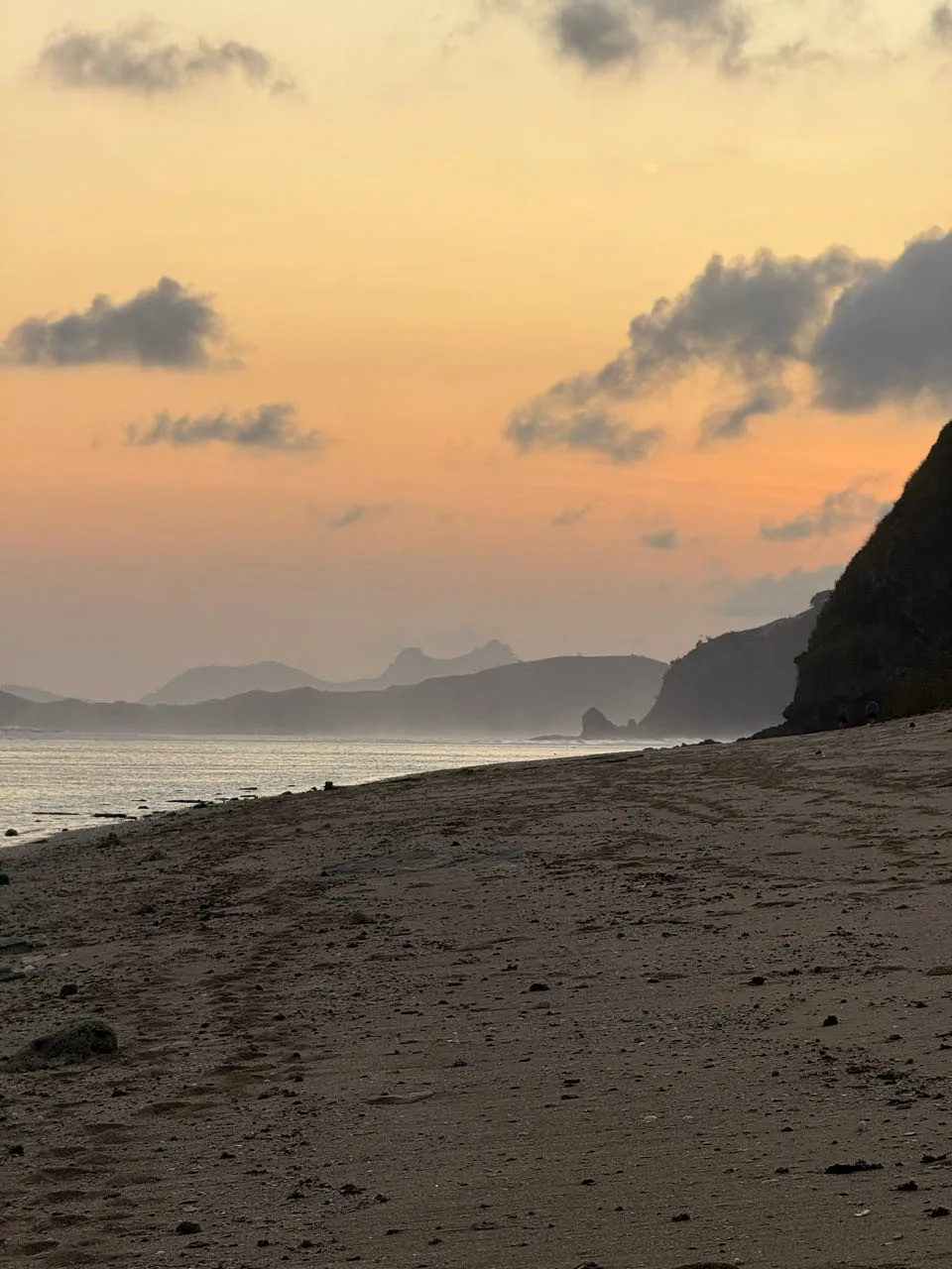 Sandstrand im orangen Abendlicht, rechts eine dunkle Felswand, in der Ferne Landzungen im Dunst