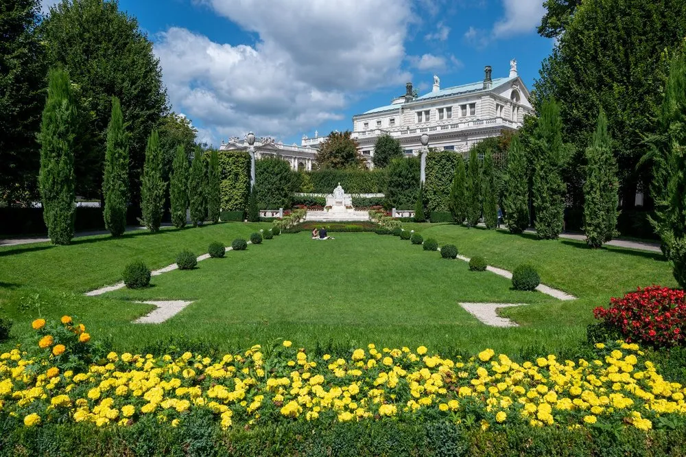 Well-kept park with lawn and a yellow-flowering bed, in the middle a white monument, behind it a building with a green roof