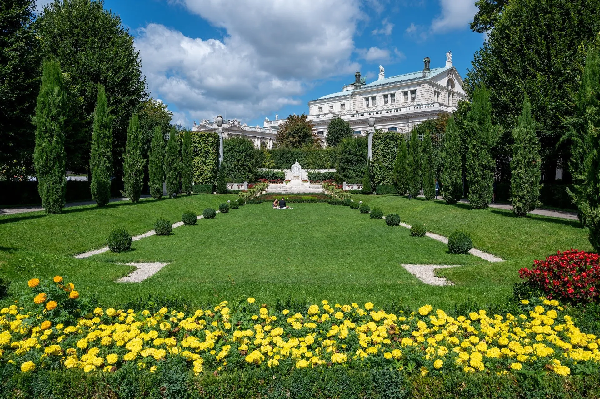 Gepflegte Parkanlage mit Rasen und gelb blühendem Beet, in der Mitte ein weisses Denkmal, dahinter ein Gebäude mit grünem Dach