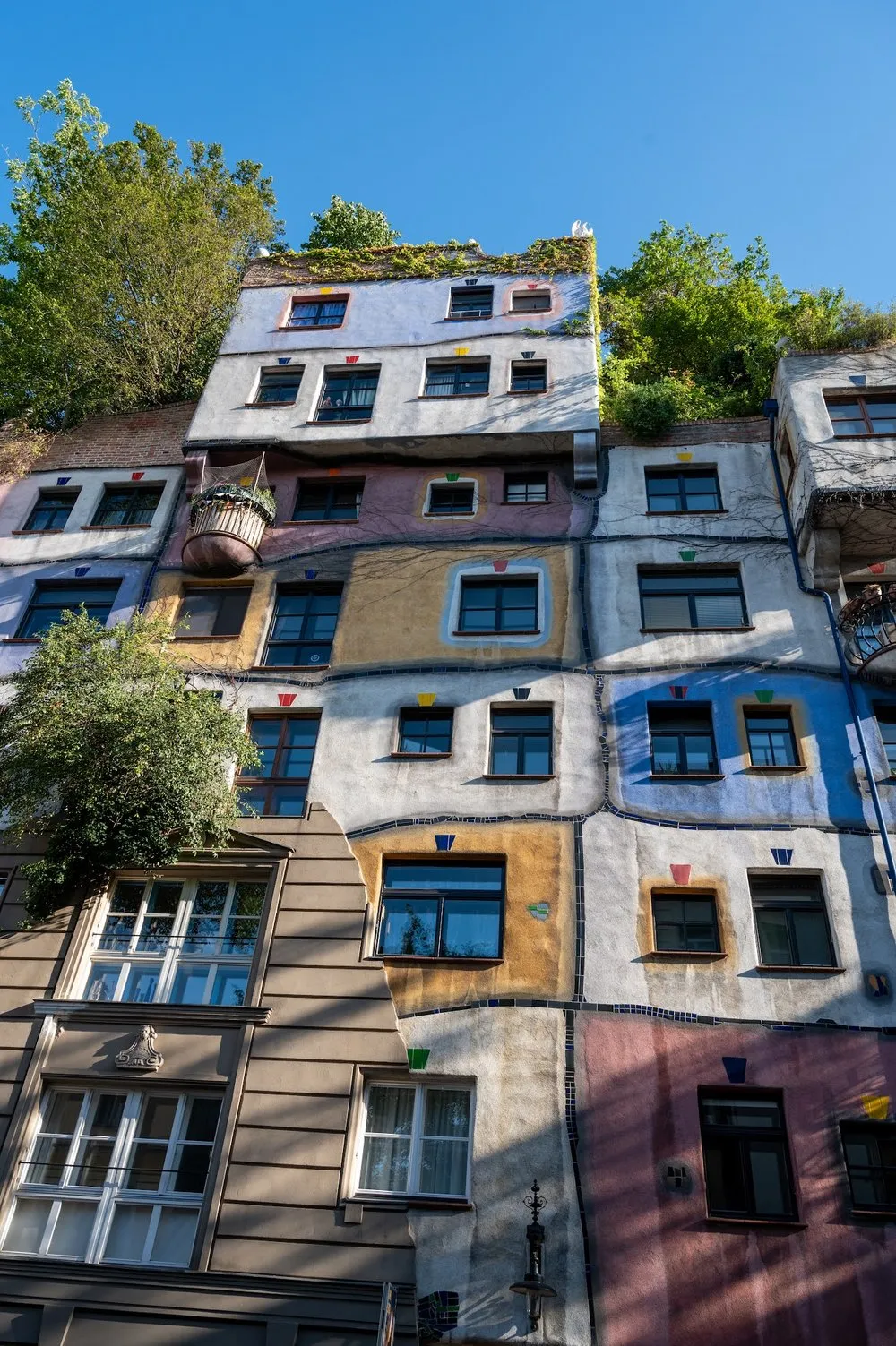 The Hundertwasser House photographed from below, at the top bushes and trees on the roof, at the bottom a pink section of façade