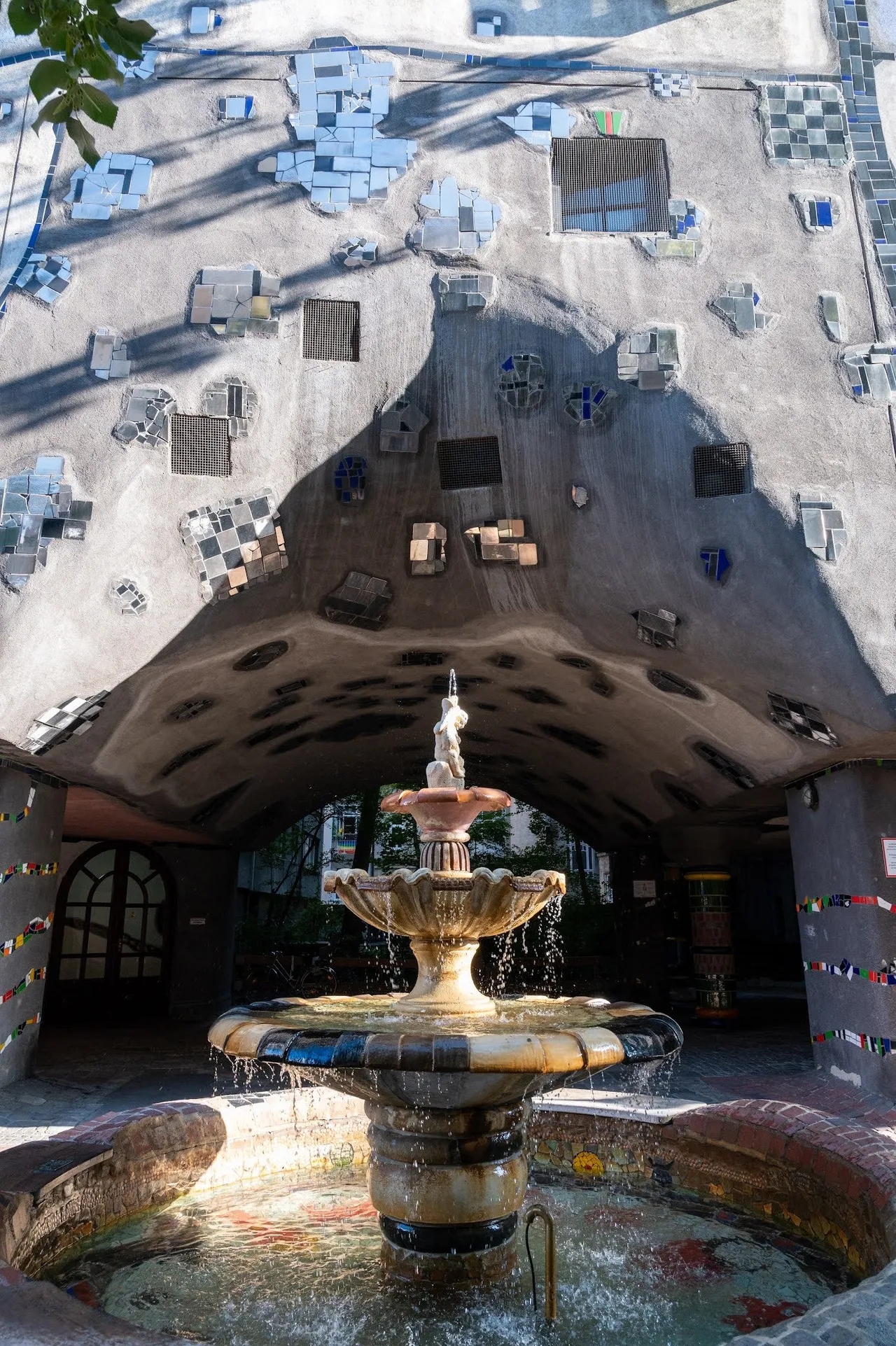 Brunnen mit gestufter Schale unter einem gewölbten Durchgang, dessen Decke mit Mosaikfeldern und kleinen Fenstern verziert ist