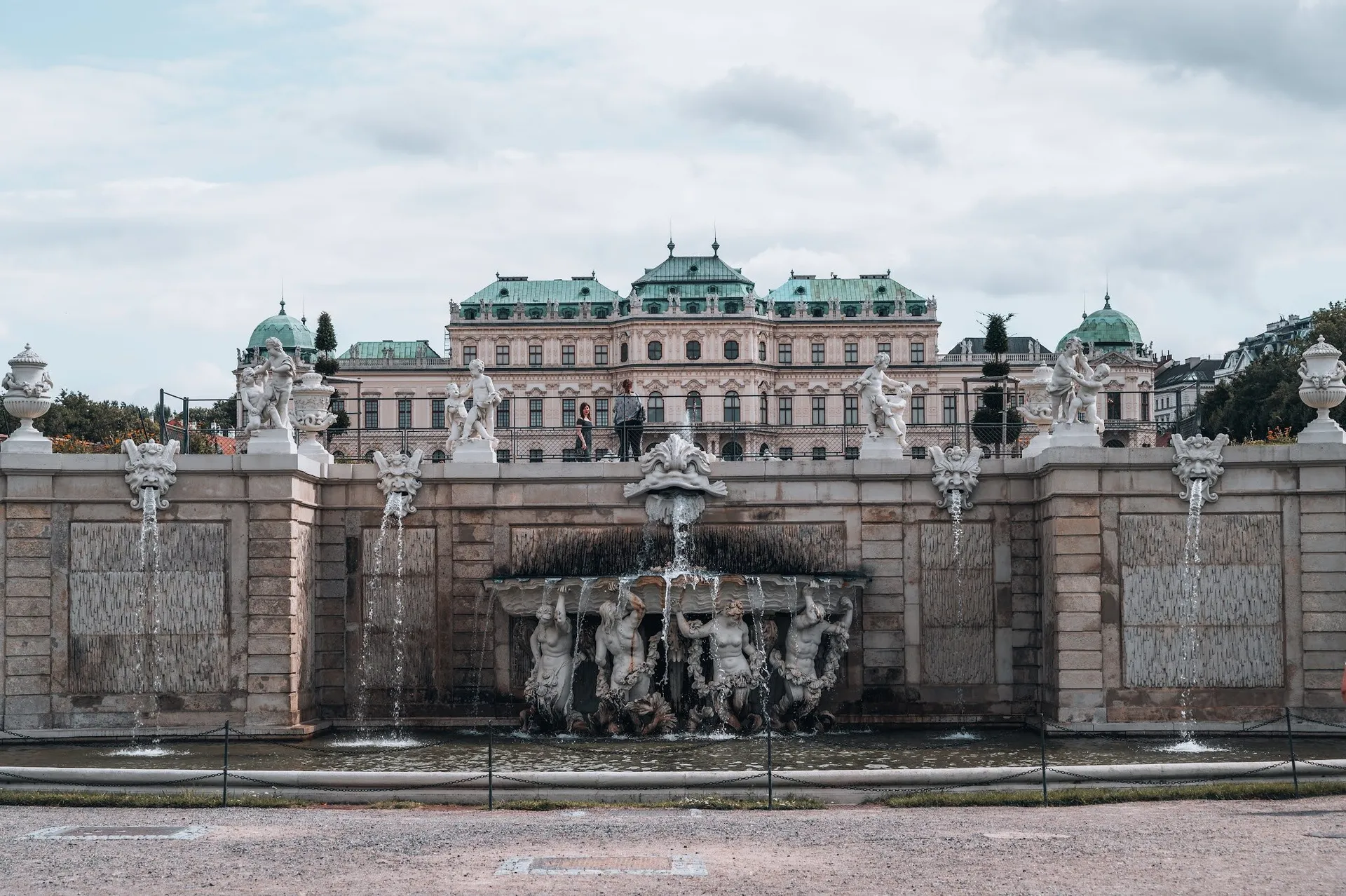 A wall with water features and statues in the garden, behind it Belvedere Palace with green domed roofs
