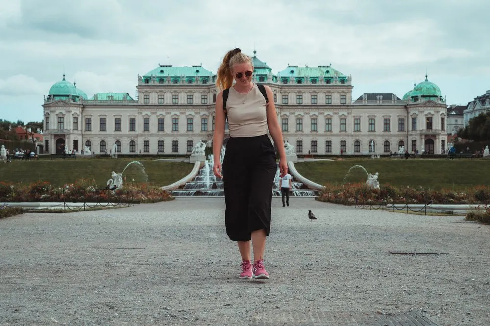 Eine Frau mit Sonnenbrille und schwarzer Hose geht über den Kiesweg, hinter ihr die Wasserspiele und das Schloss Belvedere