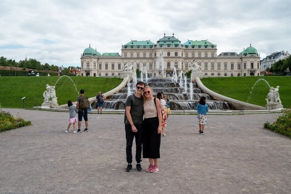 Ein Paar mit Sonnenbrillen steht vor der Wasserkaskade, dahinter das Schloss Belvedere, links und rechts weitere Besucher