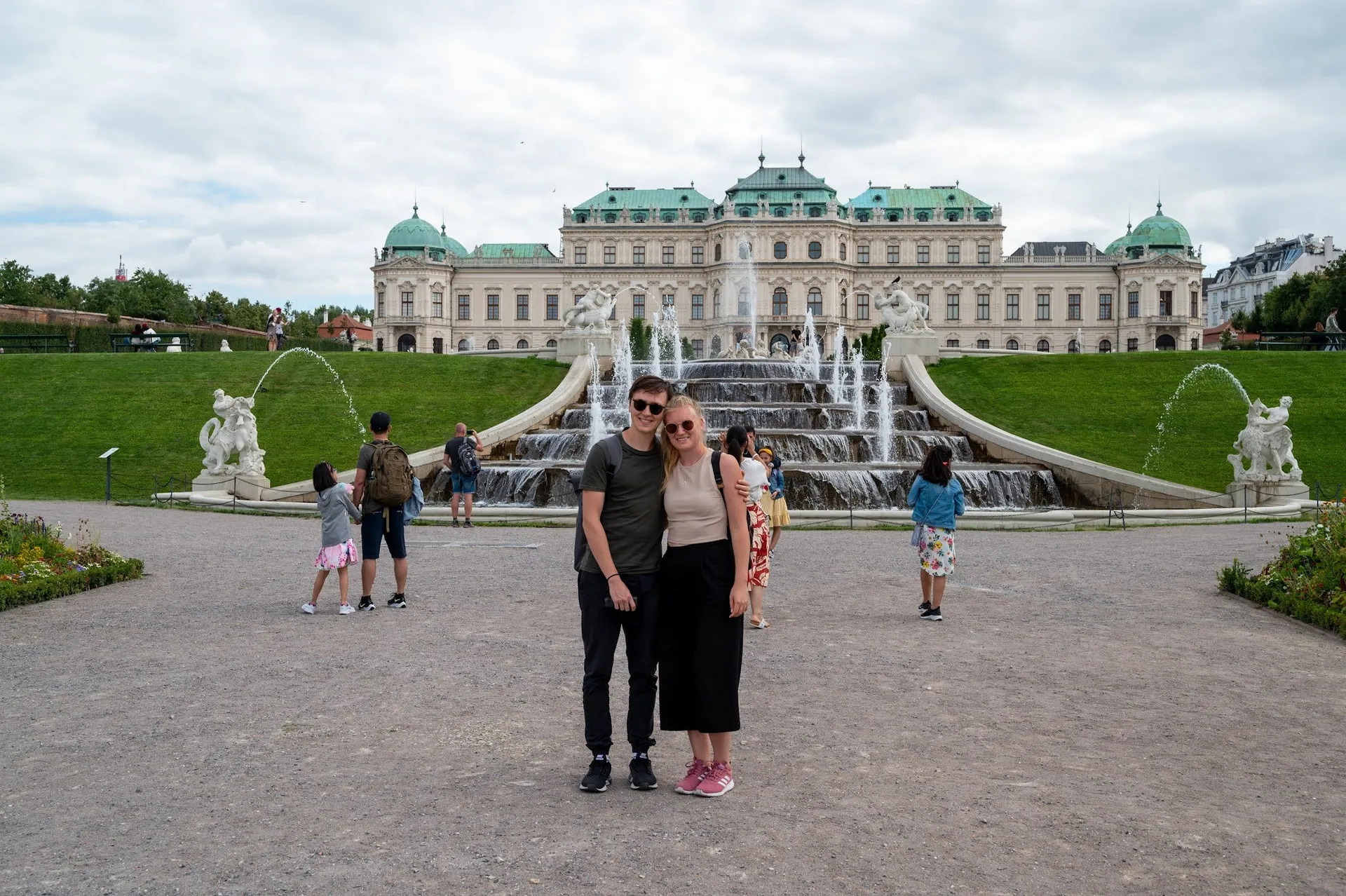 A couple in sunglasses stands in front of the water cascade, behind them Belvedere Palace, more visitors to the left and right