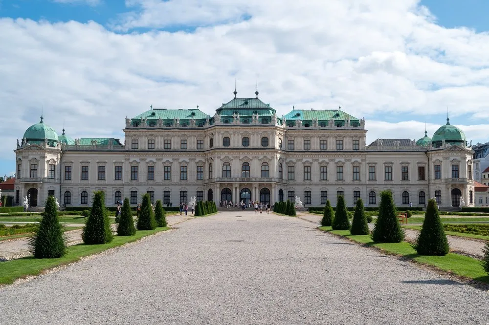 The front of Belvedere Palace with green roofs, in front of it a gravel path and ornamental trees under a blue sky with clouds