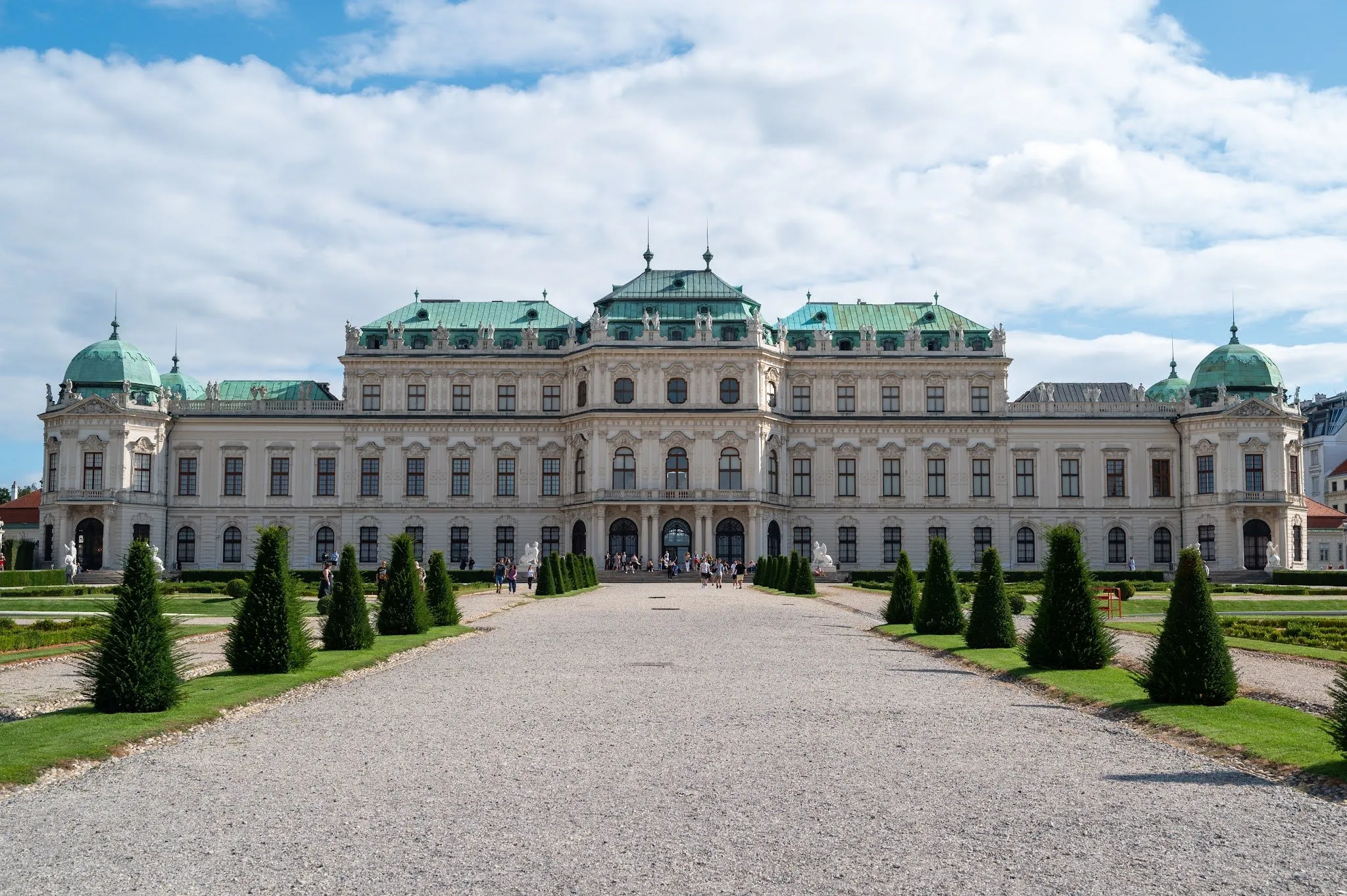 The front of Belvedere Palace with green roofs, in front of it a gravel path and ornamental trees under a blue sky with clouds
