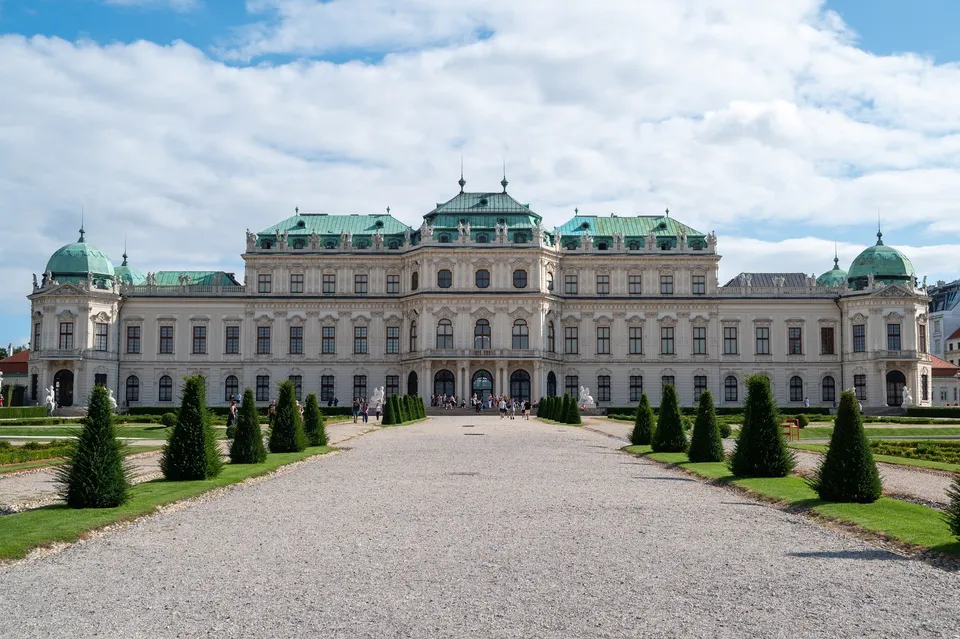Die Vorderseite von Schloss Belvedere mit grünen Dächern, davor ein Kiesweg und Zierbäume unter blauem Himmel mit Wolken