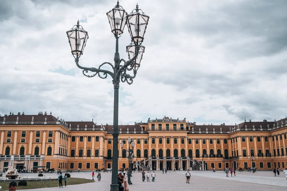 Eine verzierte Laterne mit mehreren Lampen vor der gelben Schlossfassade, dahinter Besucher auf dem Platz