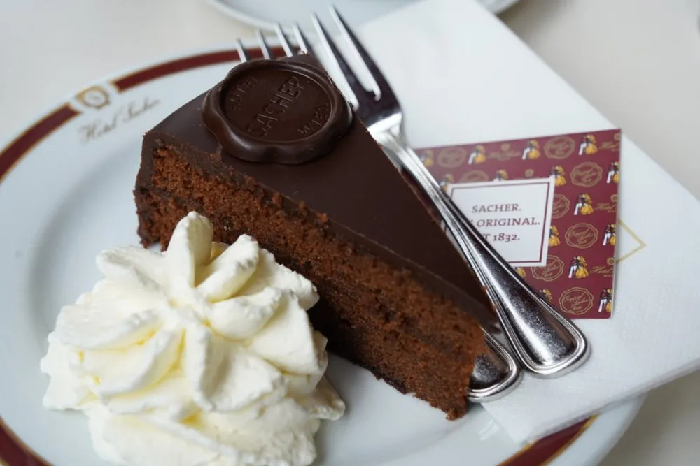 A piece of Sachertorte with chocolate glaze and a round Sacher seal, next to whipped cream and two forks on a plate