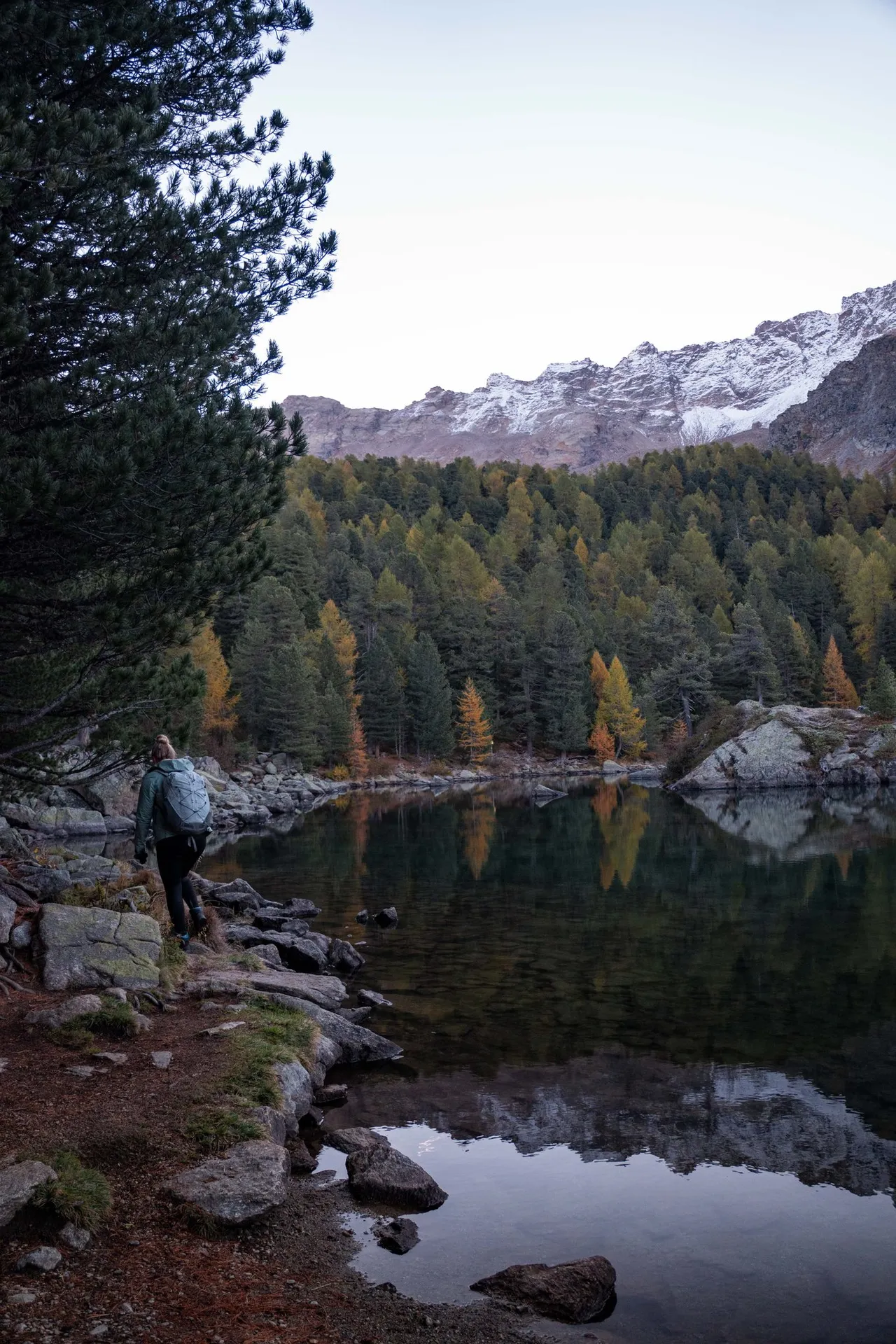 Jemand von uns geht mit Rucksack am Blockufer des Lagh da Saoseo entlang, der See liegt noch im Schatten, darüber ein frisch verschneiter Grat